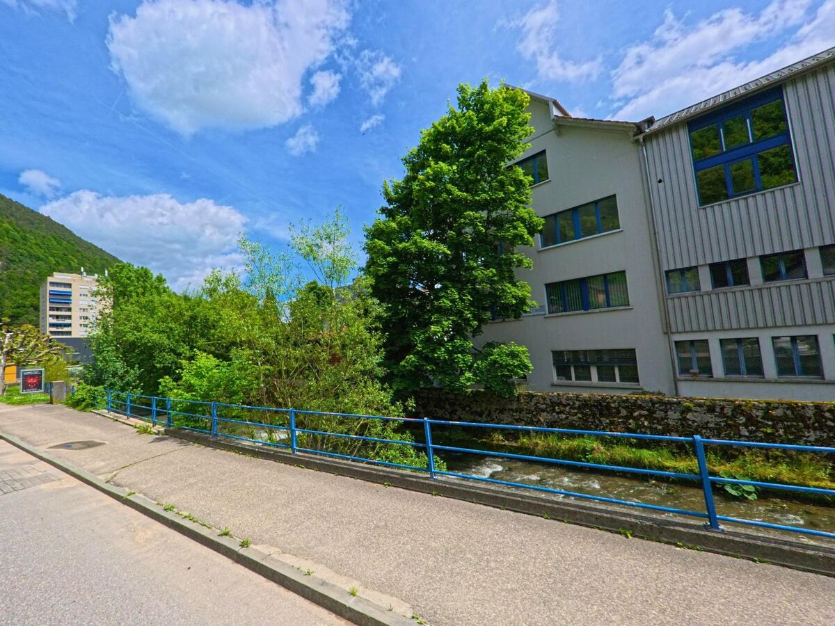 In Moutier (2740, Kanton Jura) fliesst ein schmaler Bach entlang eines Gehwegs mit blauem Geländer, daneben steht ein modernes Gebäude mit grossen Fenstern.