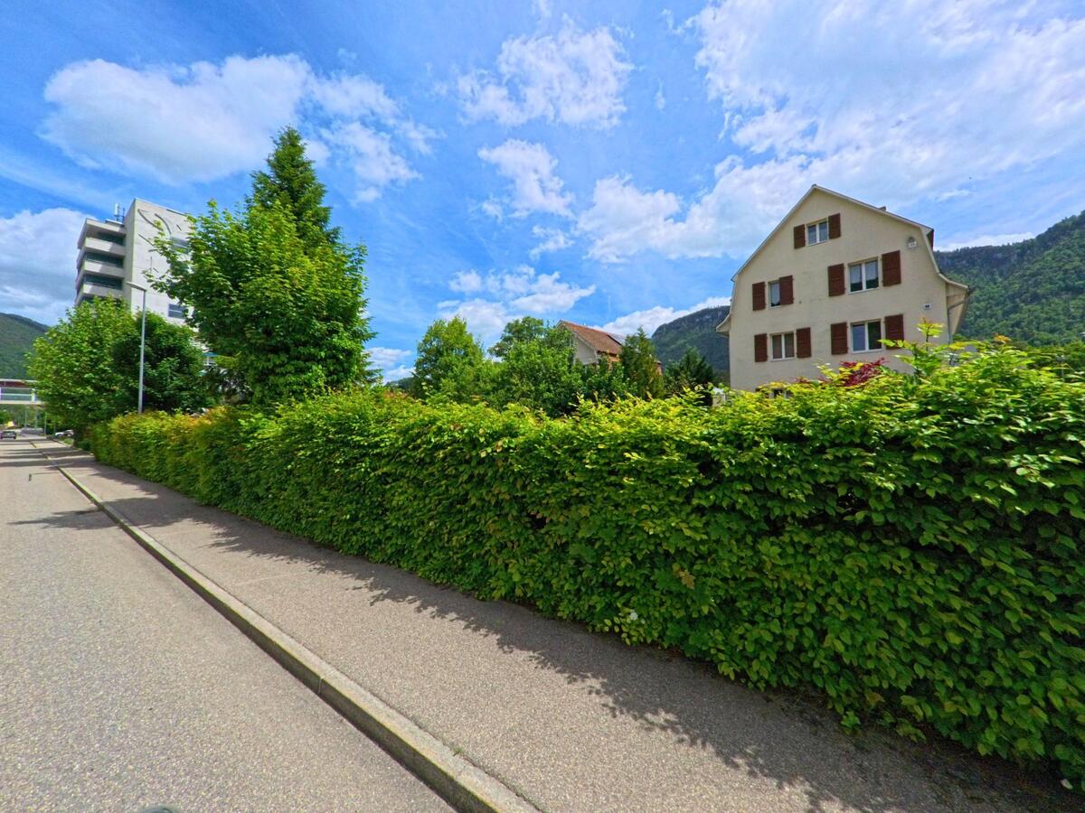 In Moutier (2740, Kanton Jura) verläuft ein Gehweg entlang einer dichten Hecke mit Blick auf Wohnhäuser und bewaldete Hügel im Hintergrund.