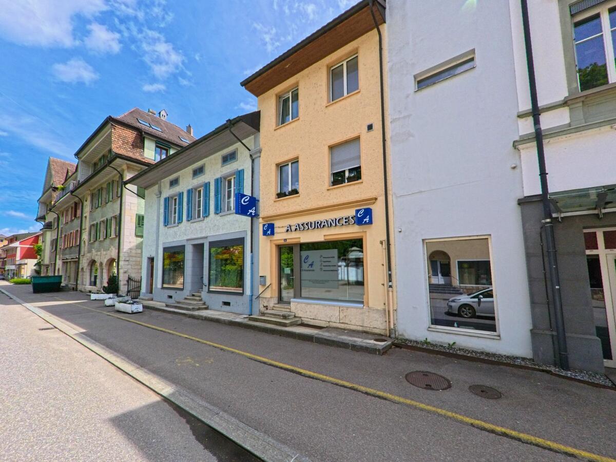 In Moutier (2740, Kanton Jura) reihen sich entlang einer schmalen Strasse farbenfrohe Geschäftshäuser mit Schaufenstern und Fensterläden.