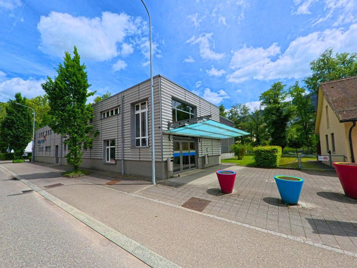 In Moutier (2740, Kanton Jura) steht ein modernes Gebäude mit gerippter Fassade und gläsernem Eingangsvordach, davor bunte Pflanzkübel auf einem gepflasterten Vorplatz.