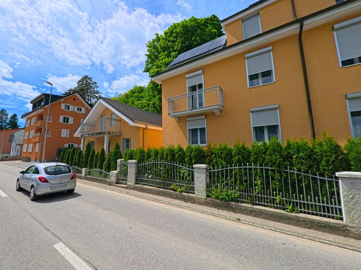 In Moutier (2740, Kanton Jura) säumen moderne Wohnhäuser mit orangefarbener Fassade und gepflegter Einfriedung eine ruhige Strasse, ein Auto fährt daran vorbei.
