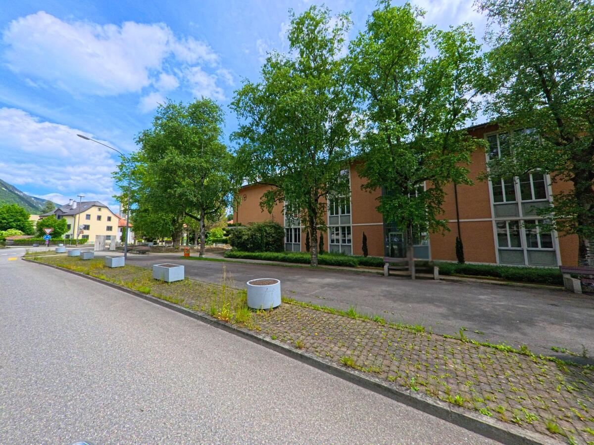 In Moutier (2740, Kanton Jura) liegt ein flaches Verwaltungs- oder Schulgebäude hinter einem Platz mit Bäumen, Bänken und Pflanzkübeln.