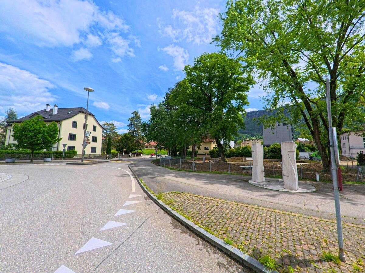 In Moutier (2740, Kanton Jura) zeigt sich eine verkehrsberuhigte Strassenszene mit moderner Skulptur im Vordergrund und Wohnhäusern zwischen Bäumen.