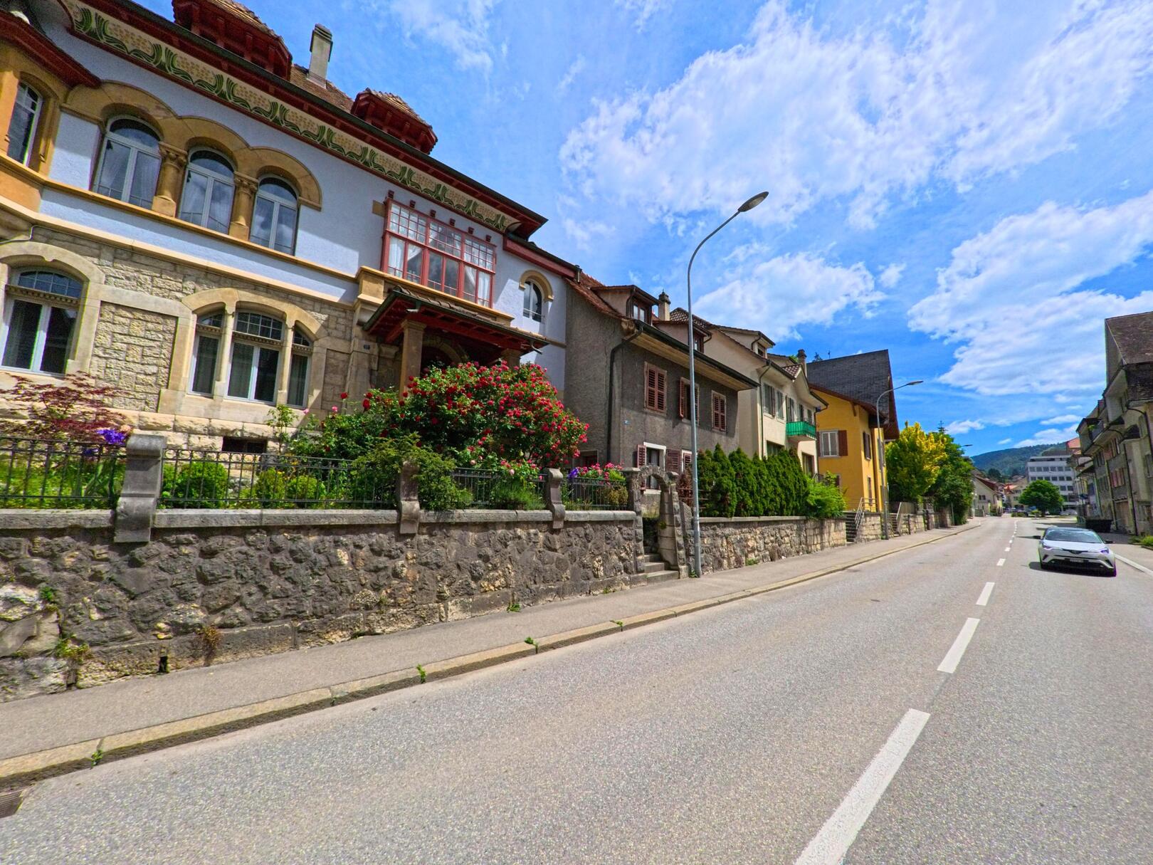 In Moutier (2740, Kanton Bern) säumen elegante Stadtvillen mit gepflegten Vorgärten eine breite Strasse unter sommerlichem Himmel.