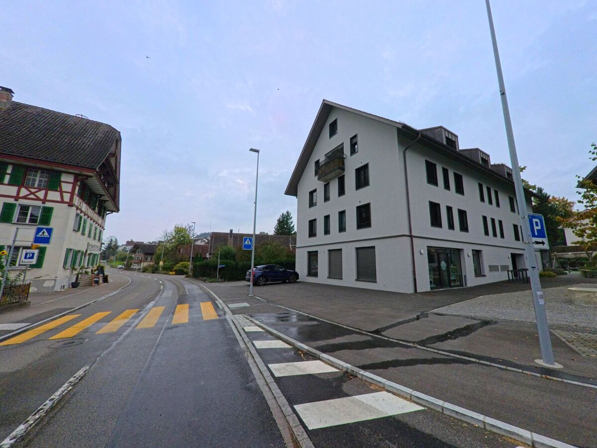 In Mühlau (5642, Kanton Aargau) verläuft eine Hauptstrasse mit Fussgängerstreifen zwischen einem traditionellen Fachwerkhaus und einem modernen Wohngebäude.