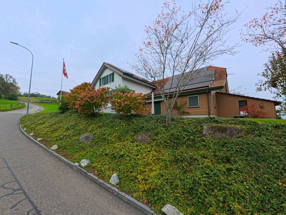 Ein Einfamilienhaus mit Solarpanels und gepflegtem Vorgarten steht in Mühlau (5642, Kanton Aargau) an einer ansteigenden Strasse mit Schweizer Fahne.