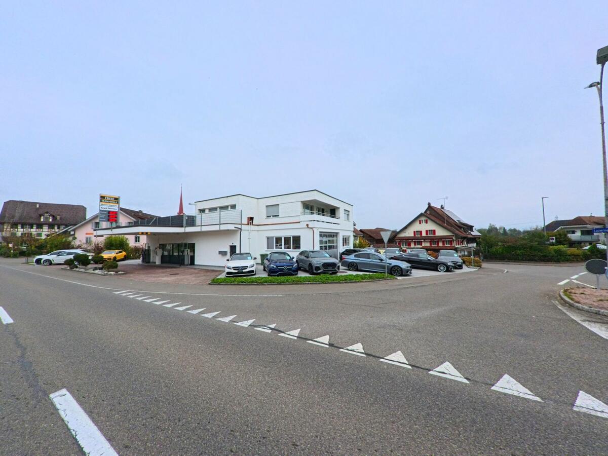 An einer Kreuzung in Mühlau (5642, Kanton Aargau) befindet sich ein modernes Gebäude mit Garagen und mehreren geparkten Autos vor traditionellen Wohnhäusern.