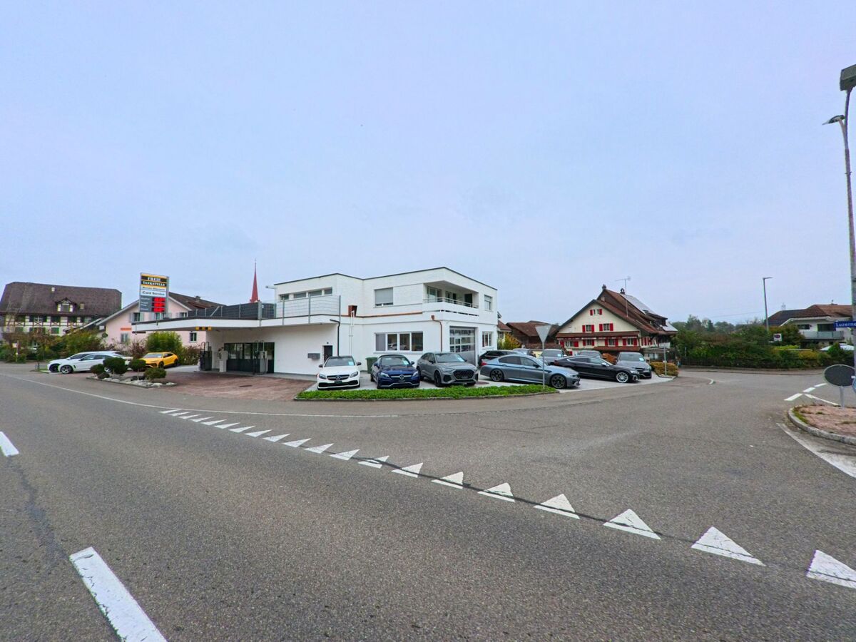 An einer Kreuzung in Mühlau (5642, Kanton Aargau) befindet sich ein modernes Gebäude mit Garagen und mehreren geparkten Autos vor traditionellen Wohnhäusern.