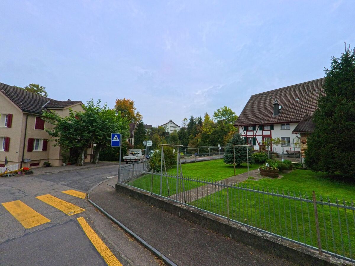 Ein Fussgängerstreifen in Mühlau (5642, Kanton Aargau) führt zu einer ruhigen Quartierstrasse mit gepflegten Gärten und traditionellen Wohnhäusern.