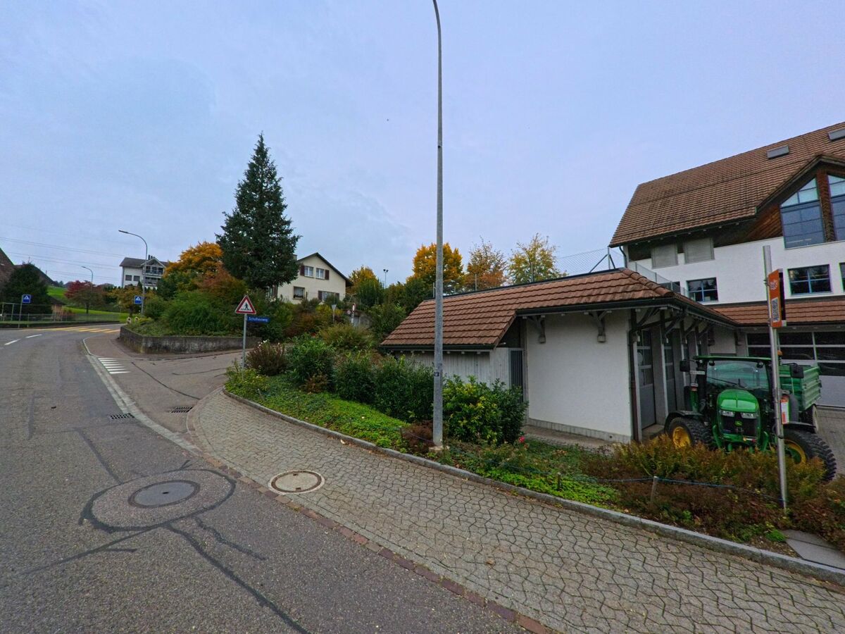 An einer Kurve in Mühlau (5642, Kanton Aargau) steht ein Gebäude mit rotem Ziegeldach und grünem Traktor neben der Strasse, umgeben von herbstlicher Vegetation.