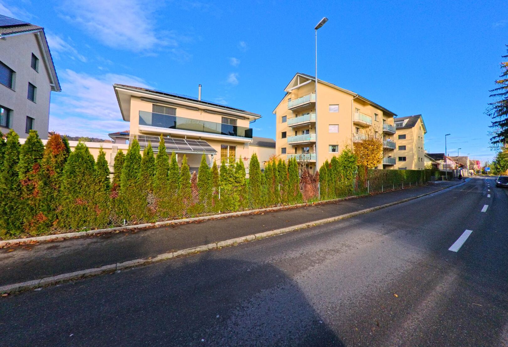 Entlang einer ruhigen Wohnstrasse in Müllheim (8555, Kanton Thurgau) stehen moderne Mehrfamilienhäuser hinter einer hohen Hecke unter blauem Himmel.