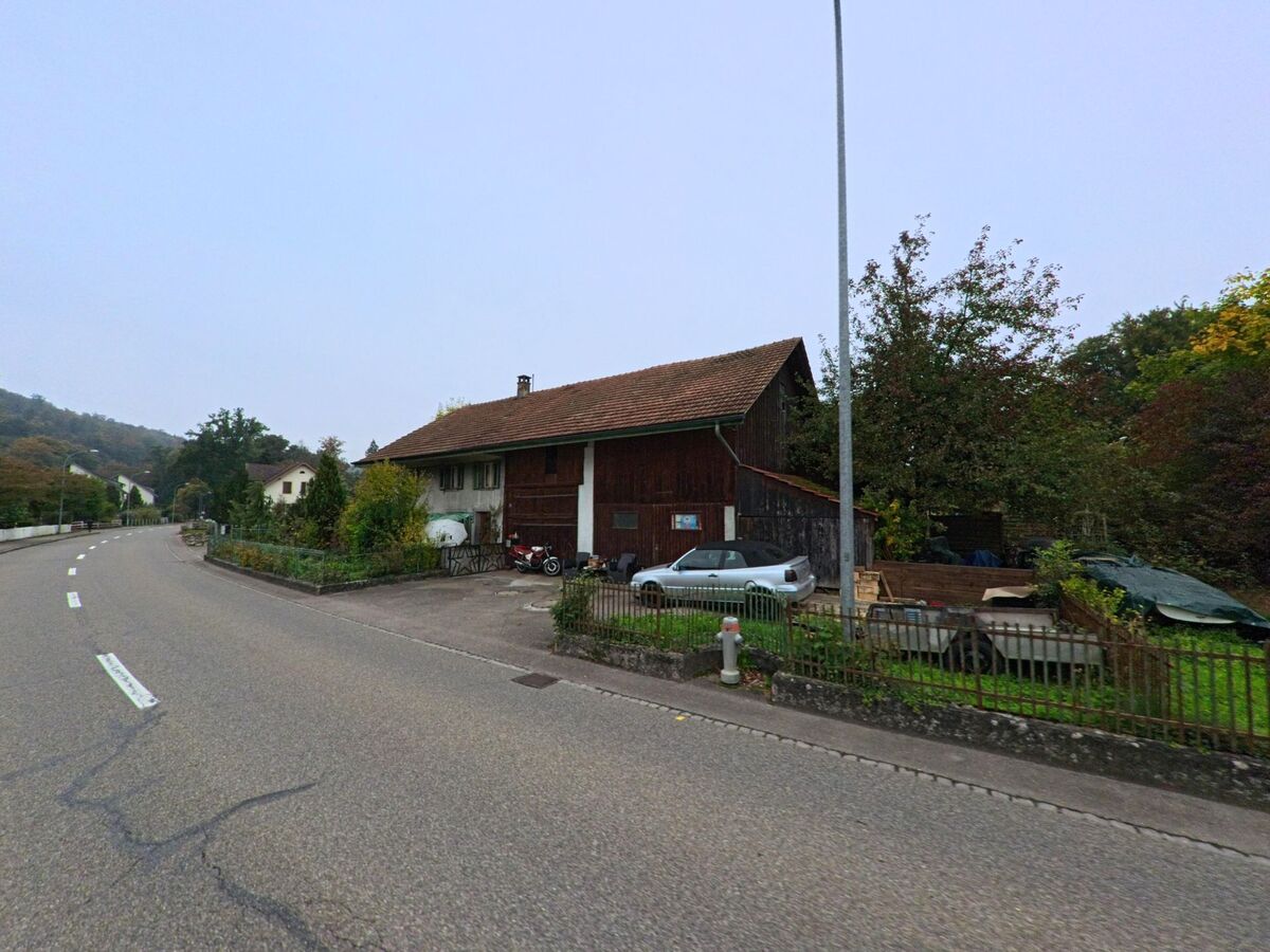 Ein älteres Bauernhaus mit Scheune und Hofeinfahrt steht an einer leicht kurvigen Strasse in Mülligen (5243, Kanton Aargau) umgeben von Bäumen und Gärten.