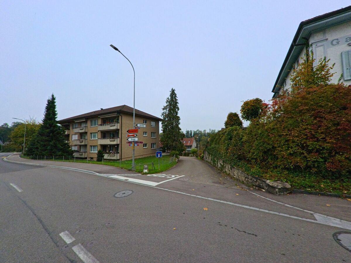 Ein Strassenkreuz in Mülligen (5243, Kanton Aargau) zeigt Wegweiser nach Brugg und Schinznach-Dorf, flankiert von Wohnhäusern und herbstlicher Vegetation.