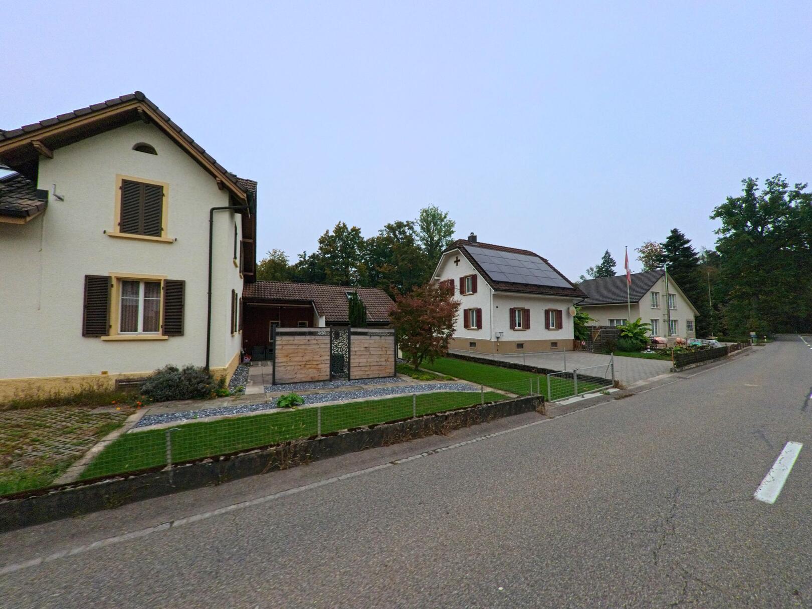 In einer ruhigen Wohnstrasse von Mülligen (5243, Kanton Aargau) stehen gepflegte Einfamilienhäuser mit Gärten und Solaranlage vor einem bewaldeten Hintergrund.