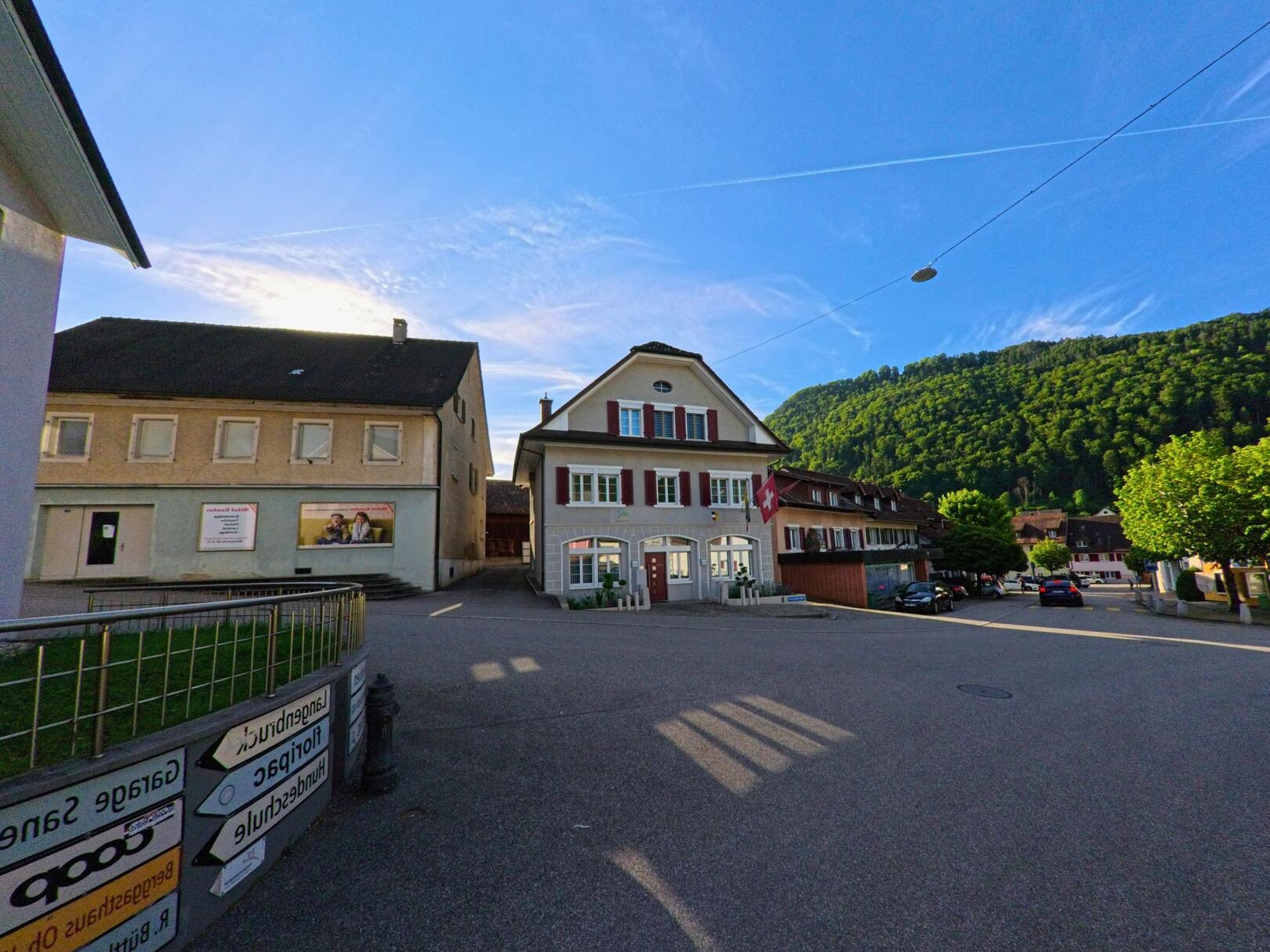 Vor dem Gemeindehaus in Mümliswil (4717, Kanton Solothurn) öffnet sich ein kleiner Platz, umgeben von traditionellen Gebäuden und bewaldeten Hügeln im Hintergrund.