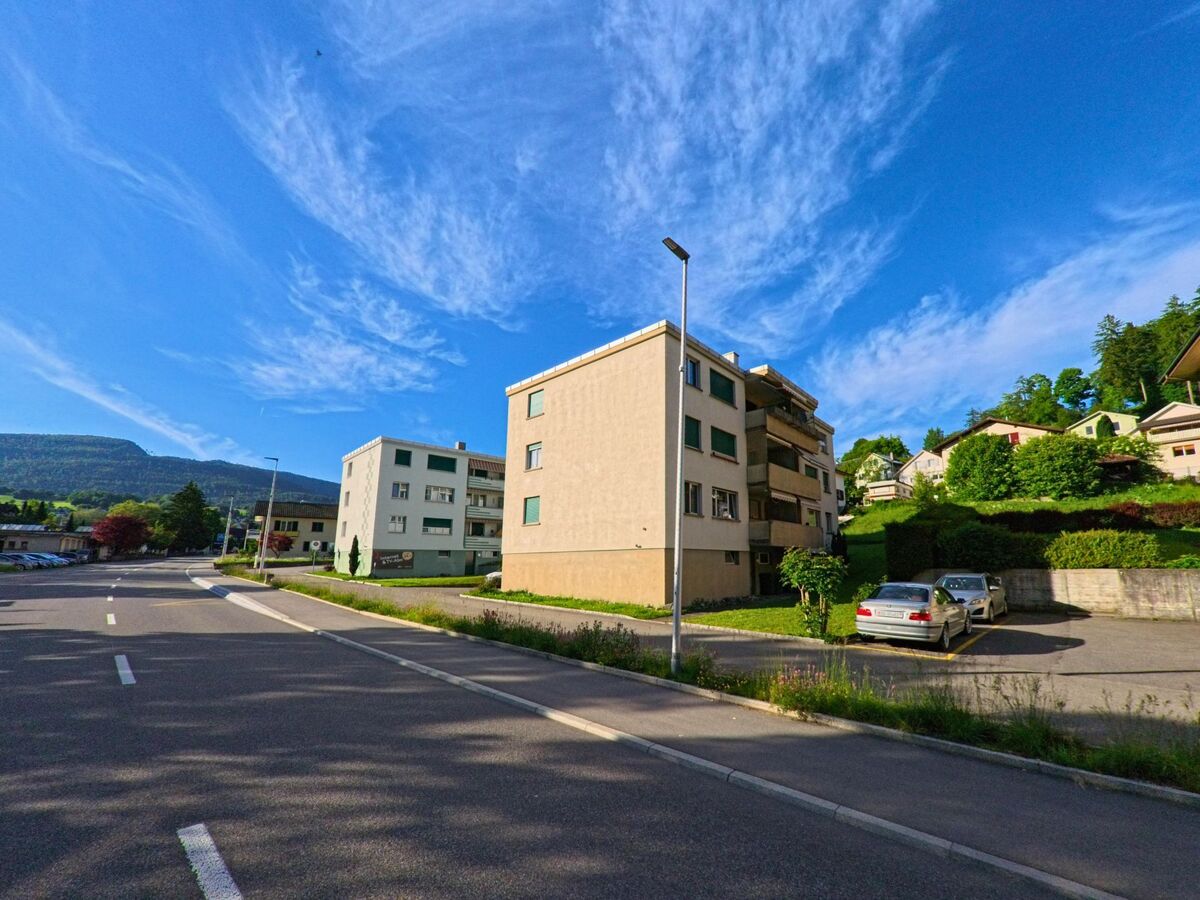 In Mümliswil (4717, Kanton Solothurn) säumen mehrere Mehrfamilienhäuser eine breite Strasse mit Parkplätzen und Blick auf bewaldete Hügel.