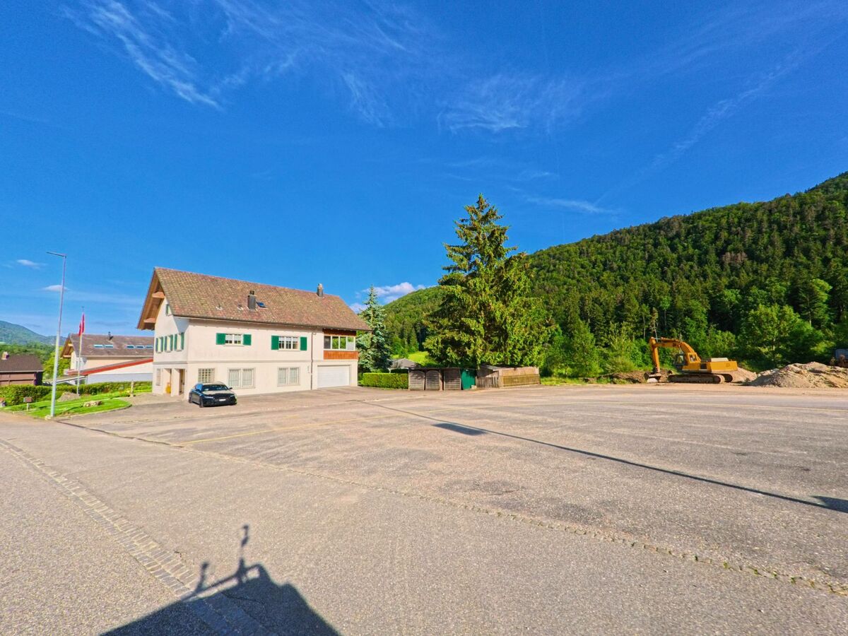 Am Rand eines grossen Parkplatzes in Mümliswil-Ramiswil (4719, Kanton Solothurn) steht ein Wohnhaus, während daneben ein Bagger auf einer Baustelle im Grünen arbeitet.