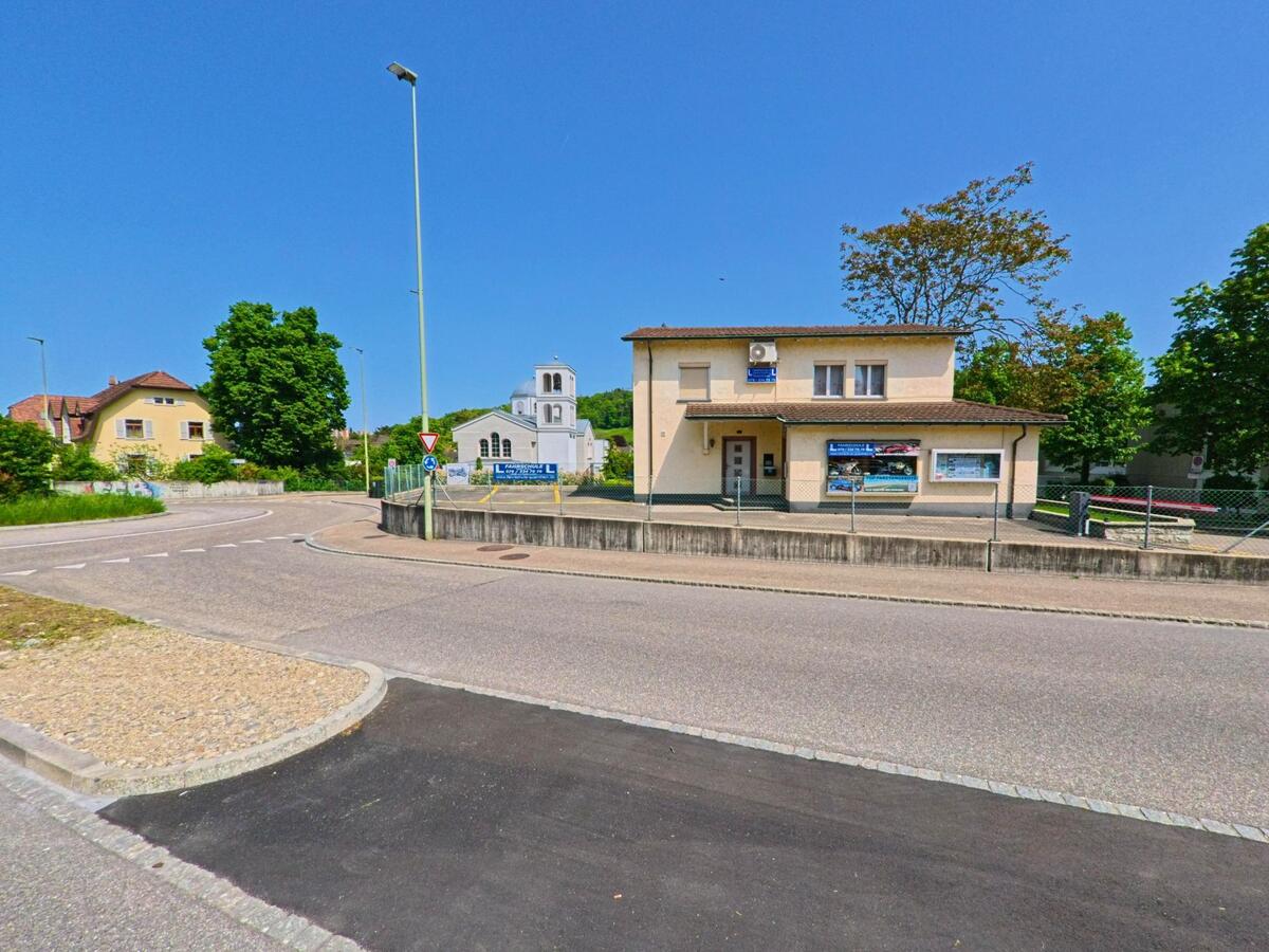 In Münchenstein (4142, Kanton Basel-Landschaft) liegt ein schlichtes Geschäftsgebäude mit angeschlossener Fahrschule an einer Strassenkreuzung, im Hintergrund ist eine weiss leuchtende Kirche zu sehen.