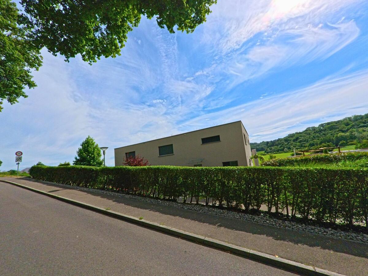 Entlang einer ruhigen Strasse in Münchenwiler (1797, Kanton Bern) steht ein modernes Wohnhaus mit Sicht auf die angrenzenden Felder und den Waldrand.