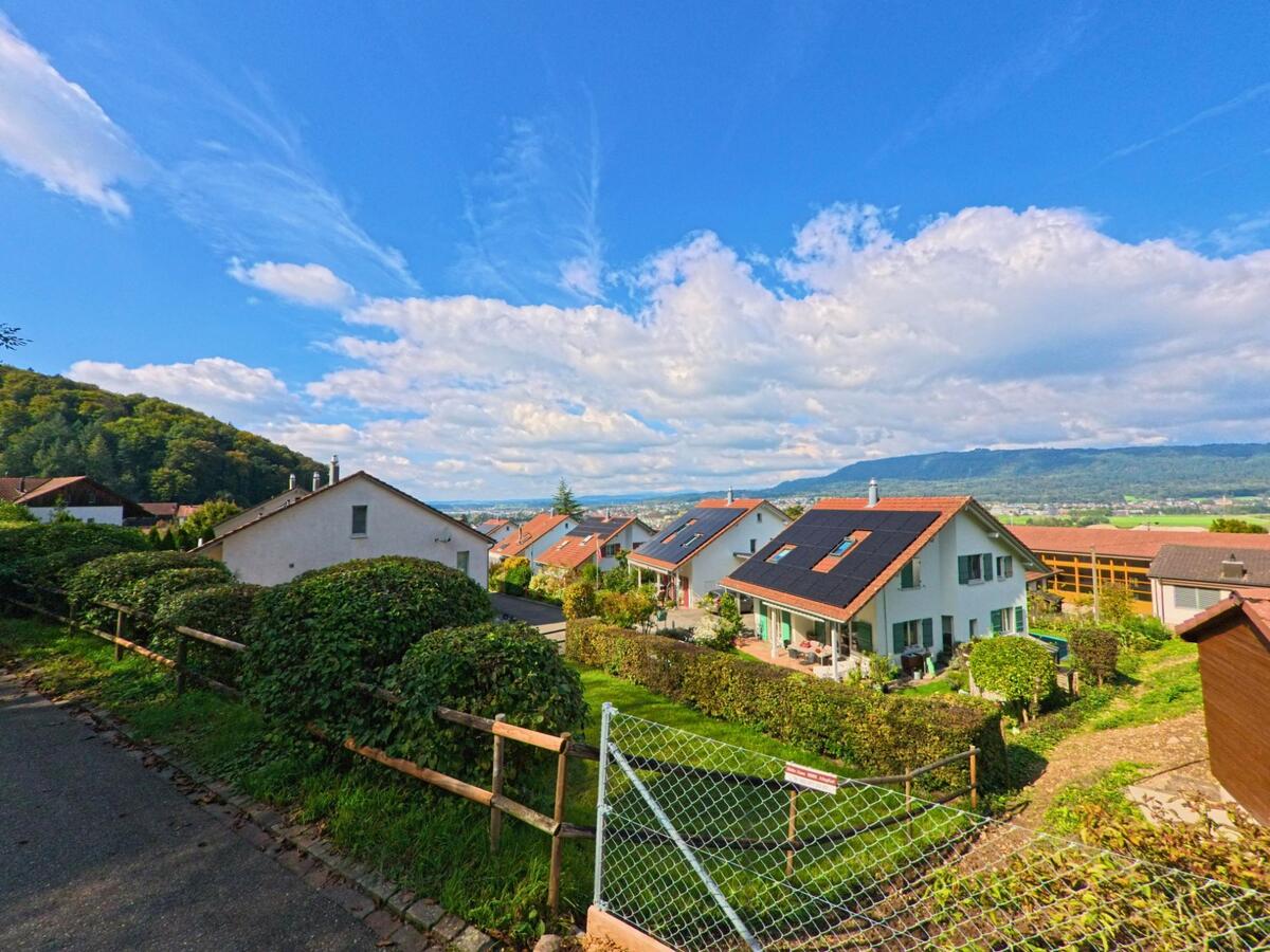 Von einem Hang in Münchwilen (4333, Kanton Aargau) bietet sich ein Blick auf moderne Wohnhaeuser mit Solardachanlagen und das darunterliegende Tal.
