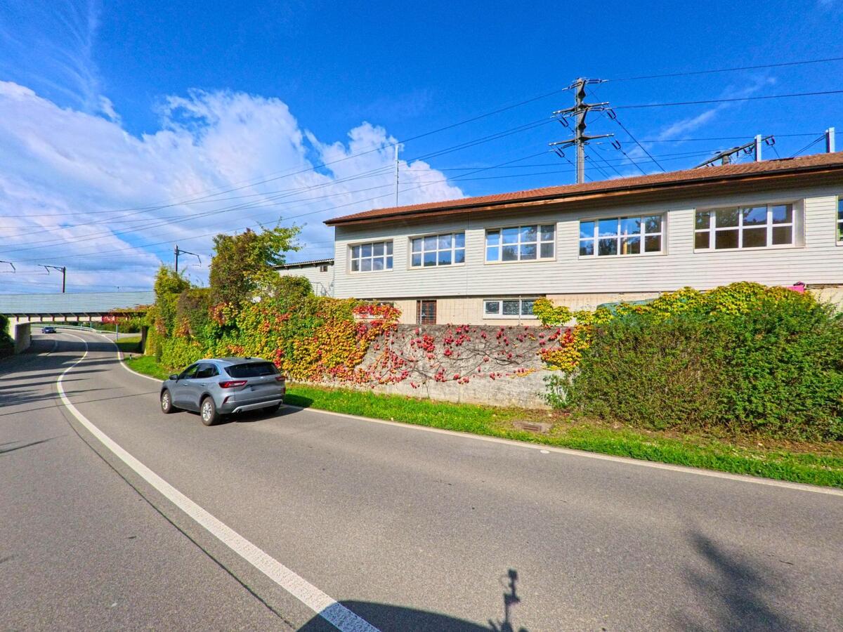 Entlang der Hauptstrasse in Münchwilen (4333, Kanton Aargau) verlaeuft eine Bahnlinie mit einem Gebaeude, dessen Fassade von farbigen Kletterpflanzen bedeckt ist.