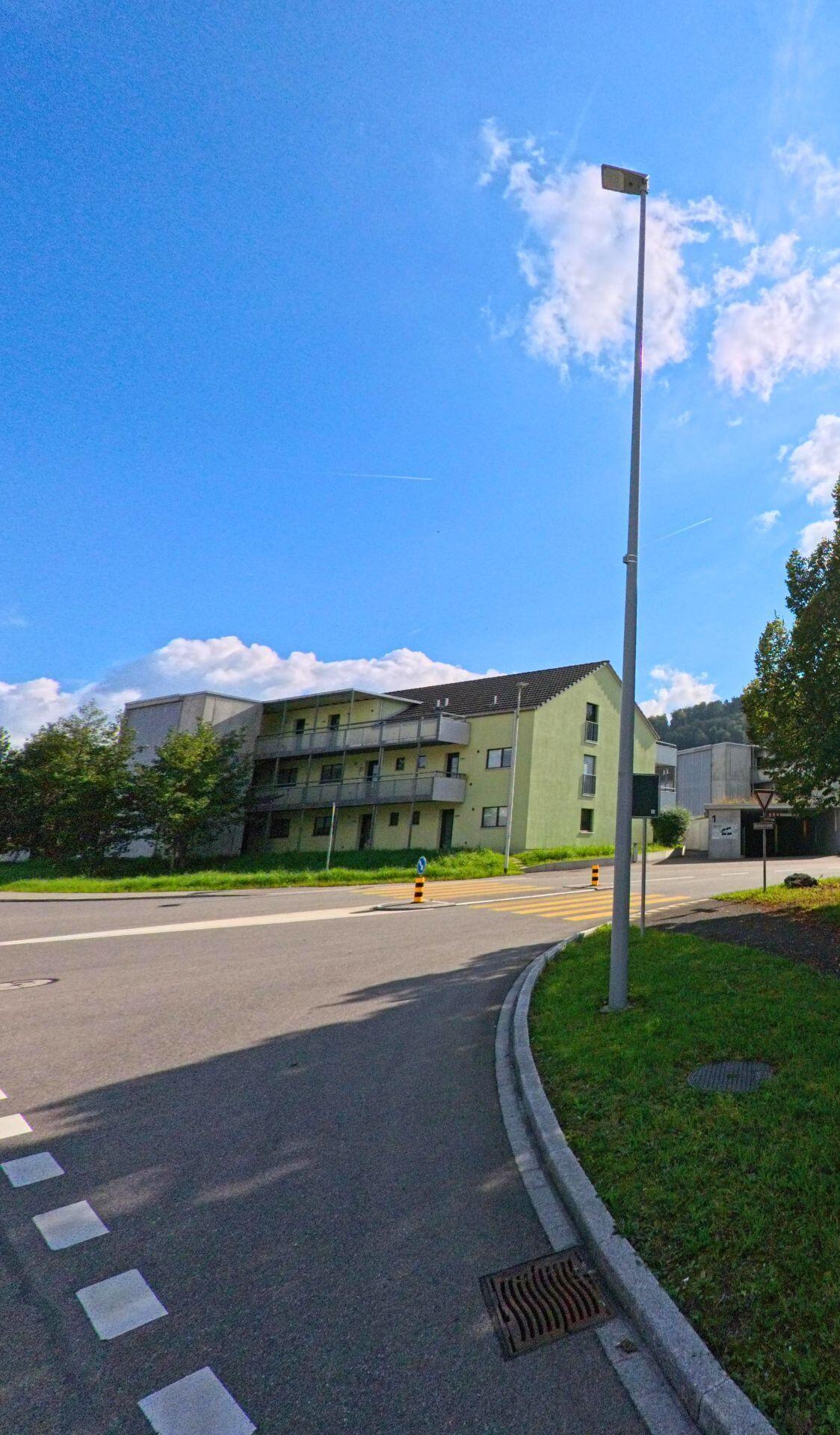Ein modernes Mehrfamilienhaus in Münchwilen (4333, Kanton Aargau) steht an einer Strassenkreuzung mit Fussgaengerstreifen und Strassenlaterne.