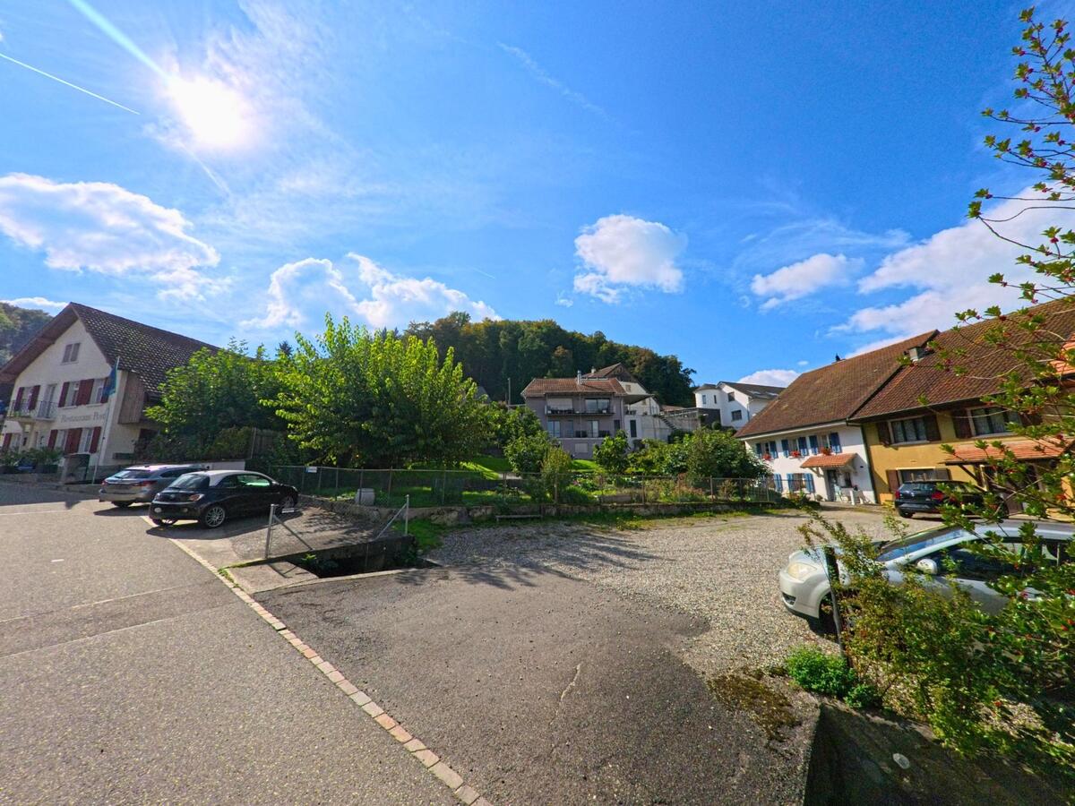 Im Ortszentrum von Münchwilen (4333, Kanton Aargau) stehen traditionelle Wohnhaeuser mit Parkplaetzen und Baumen unter einem sonnigen Himmel.