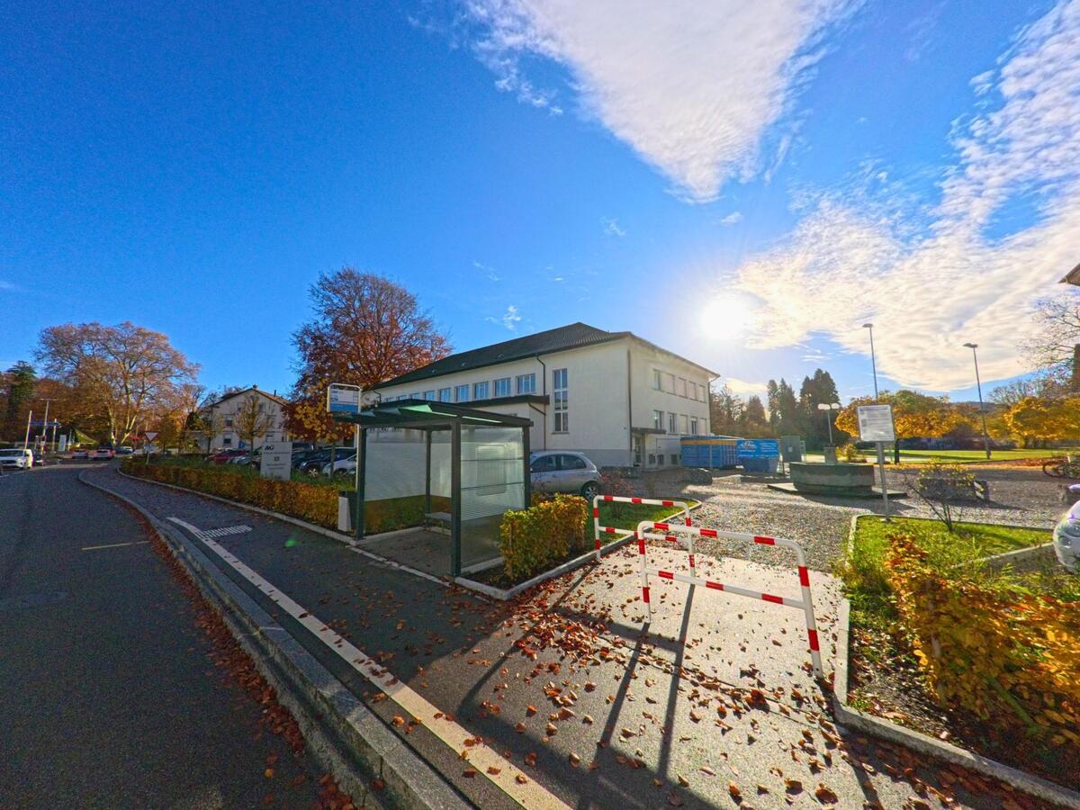 Bei einer Bushaltestelle in Münchwilen (9542, Kanton Thurgau) stehen Wartehäuschen und Absperrgitter neben einem Parkplatz vor einem hellen Gebäude im Gegenlicht der Sonne.