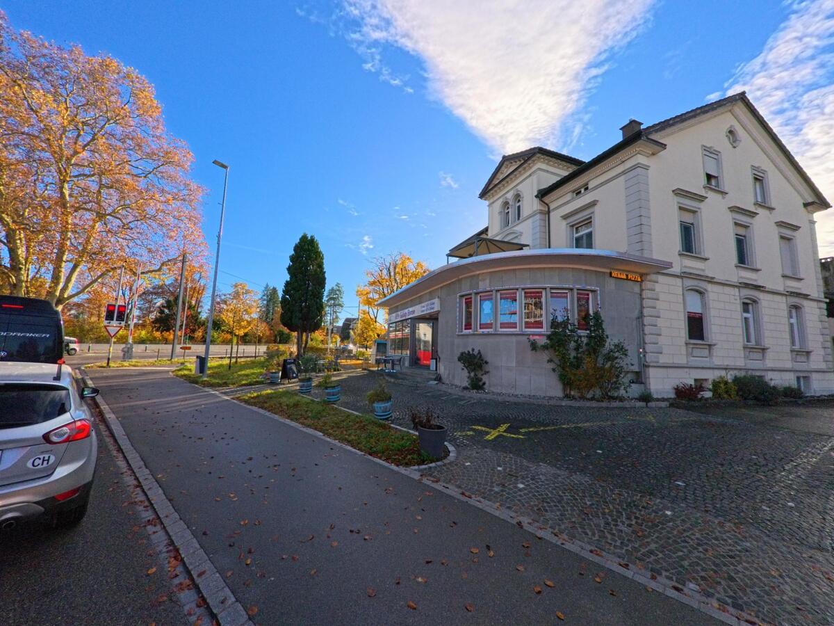 An einer Strassenecke in Münchwilen (9542, Kanton Thurgau) steht ein markantes Eckgebäude mit Ladenfront, daneben säumen Herbstbäume und Ampeln die Kreuzung.