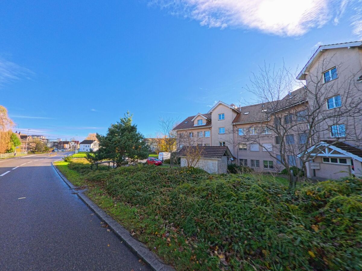 Am Rand einer Wohnstrasse in Münchwilen (9542, Kanton Thurgau) stehen Mehrfamilienhäuser hinter Hecken und Bäumen im hellen Tageslicht.