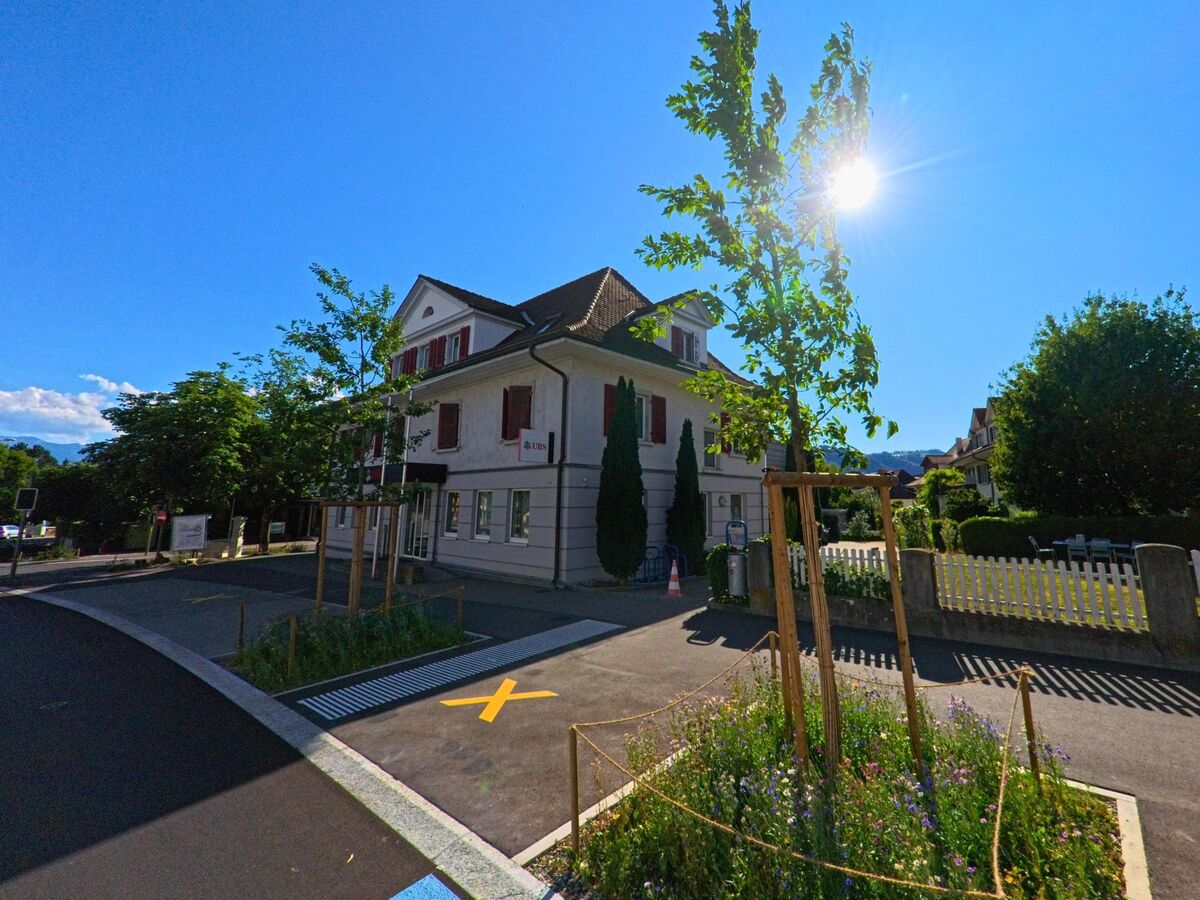 In Münsingen (3110, Kanton Bern) steht ein helles Gebäude mit roten Fensterläden und UBS-Filiale an einer ruhigen Strasse, eingerahmt von junger Bepflanzung und Sonnenschein.