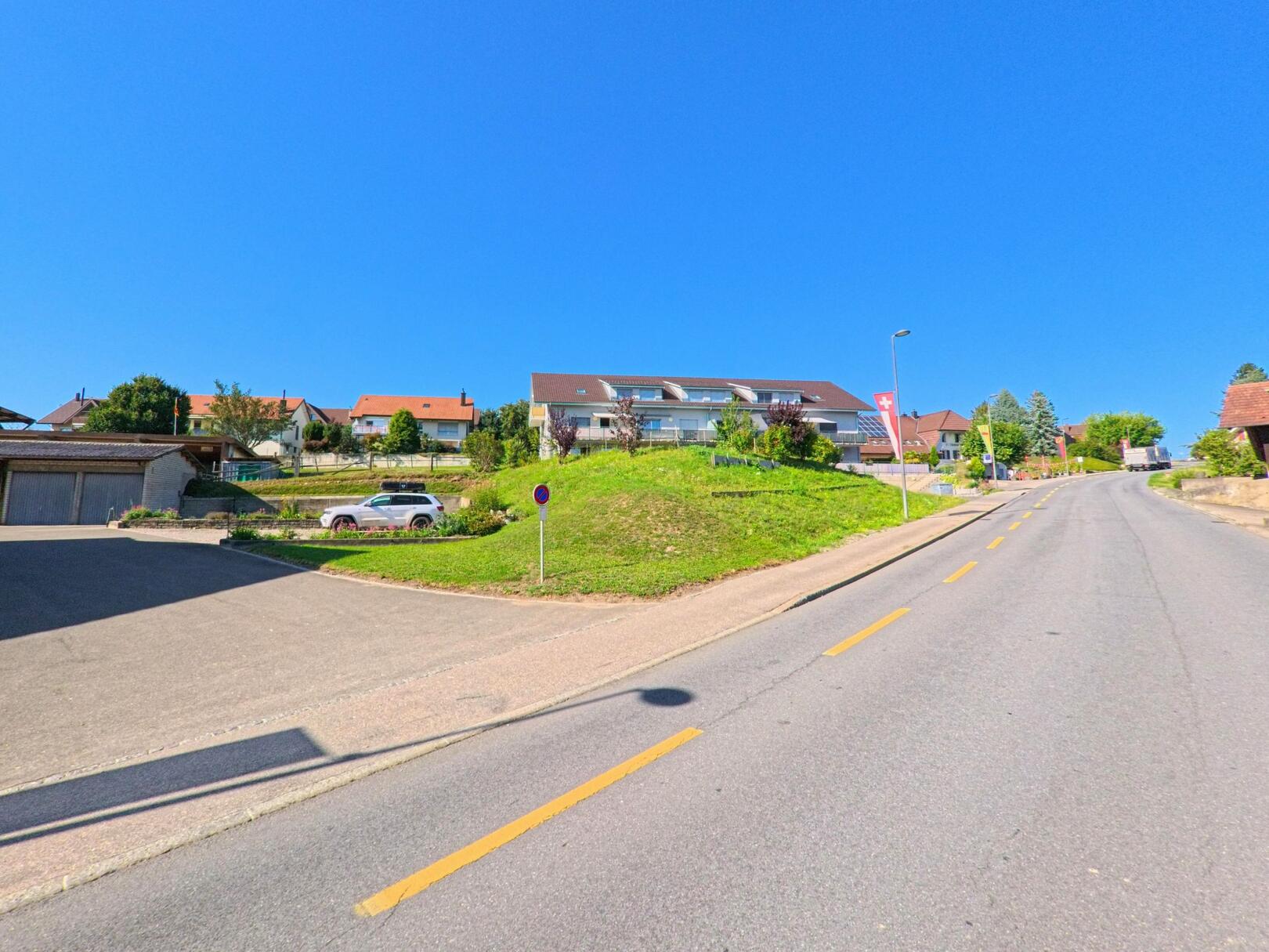 In Müntschemier (3225, Kanton Bern) steigt eine Strasse leicht an und wird von Wohnhäusern mit roten Dächern und modernen Gebäuden gesäumt.