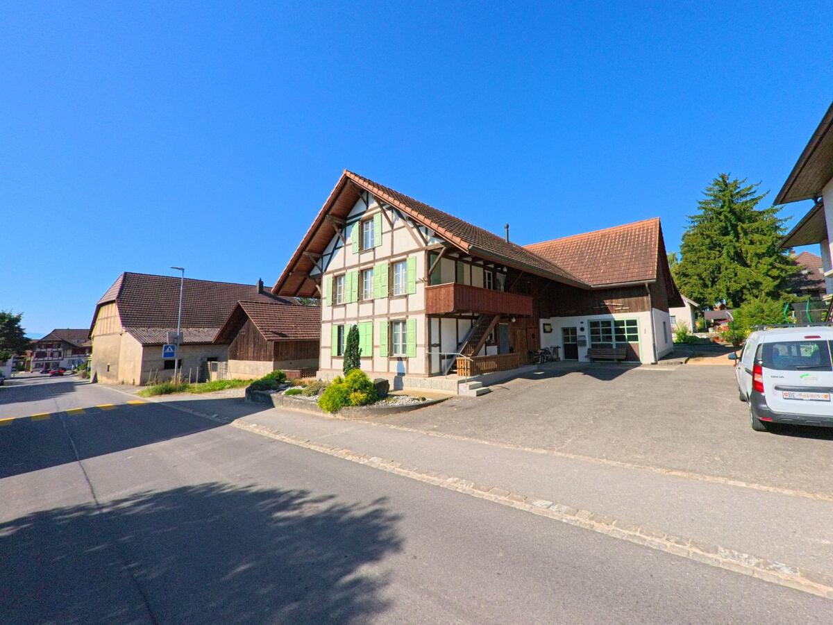 In Müntschemier (3225, Kanton Bern) steht ein traditionelles Bauernhaus mit grünen Fensterläden und grossem Vorplatz an einer Dorfstrasse.