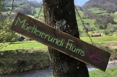 Ein Holzschild mit der Aufschrift Muettererundi haengt an einem Baum in Flums (8890, Kanton St Gallen) vor einer gruenen Huegellandschaft.