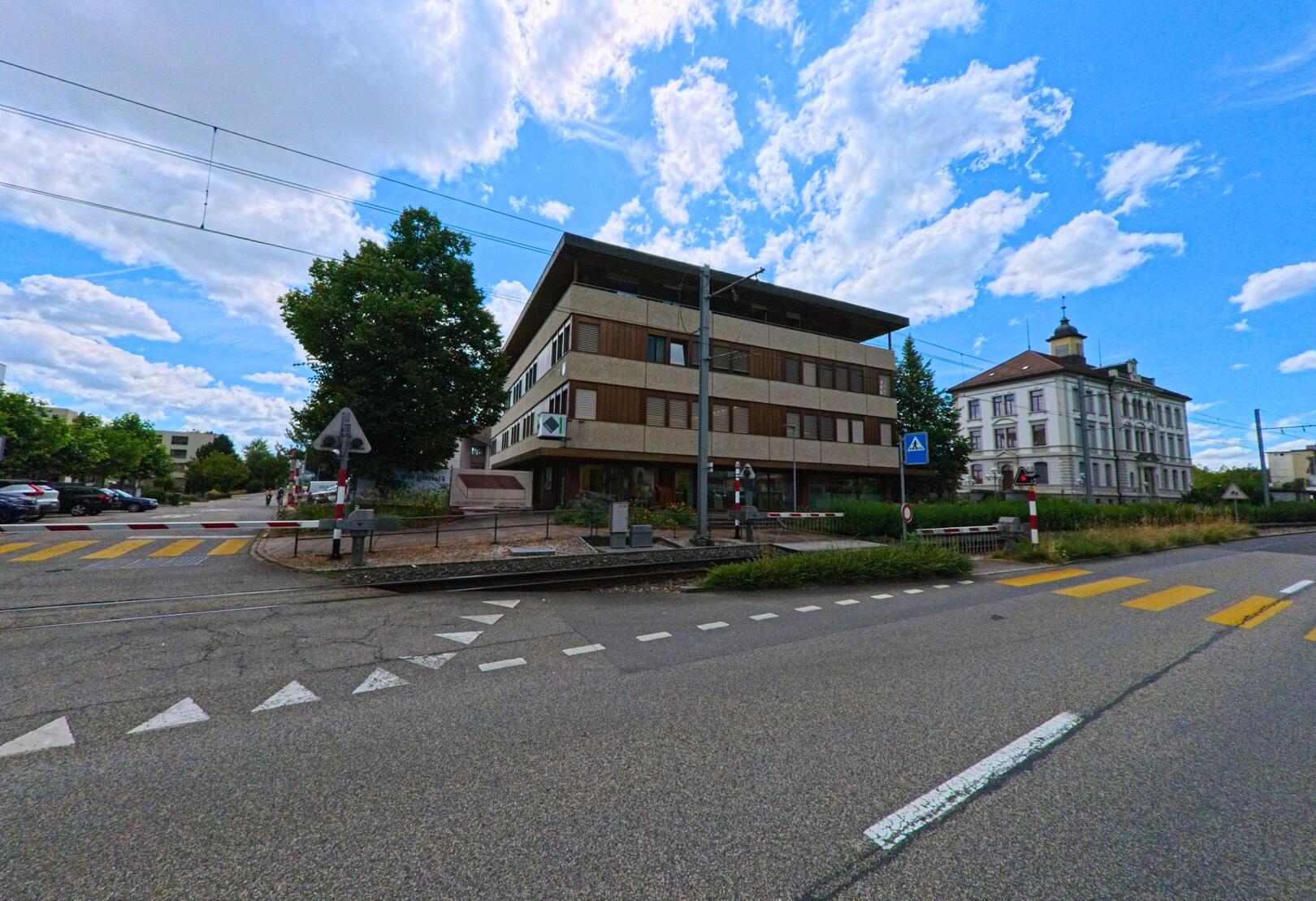 In Muhen (5037, Kanton Aargau) steht ein modernes Verwaltungsgebaeude direkt an einem Bahnuebergang, gegenueber befindet sich ein historisches Gebaeude mit Turmaufsatz.