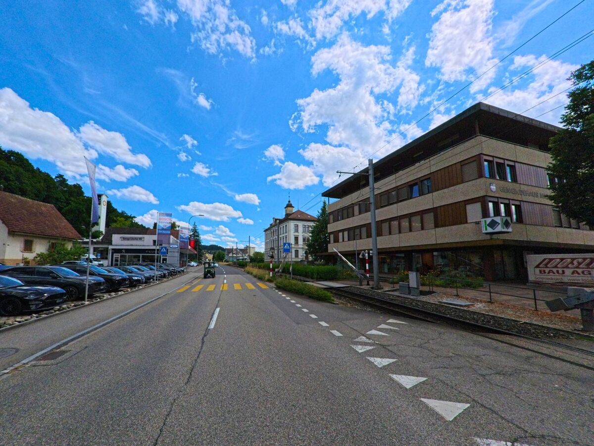 In Muhen (5037, Kanton Aargau) befinden sich ein modernes Gemeindehaus, ein Autohaendler und ein Fussgaengerstreifen entlang einer Hauptstrasse mit Bahnuebergang.
