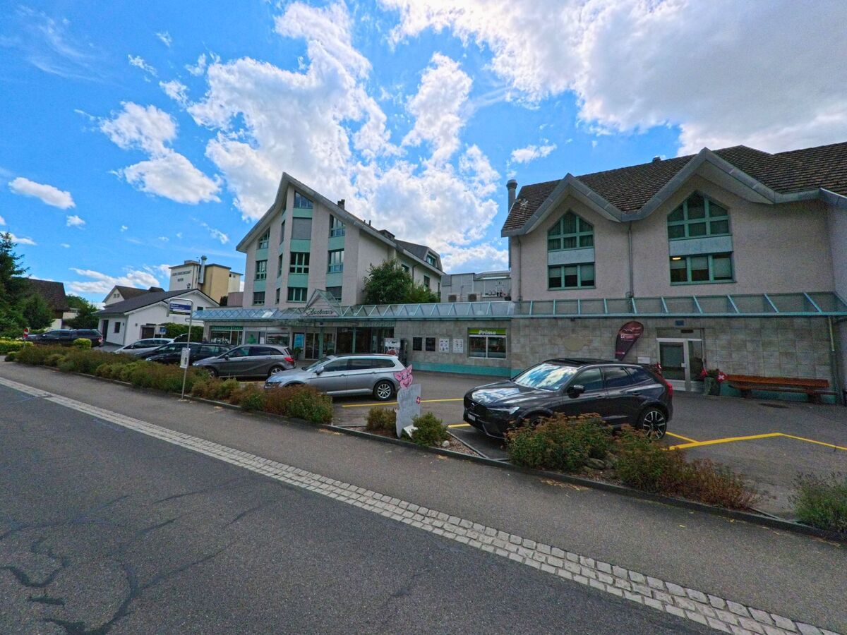 In Muhen (5037, Kanton Aargau) befindet sich ein modernes Gebaeudekomplex mit Ladenzeile und Arztpraxis direkt an einer Strasse mit Parkplaetzen.