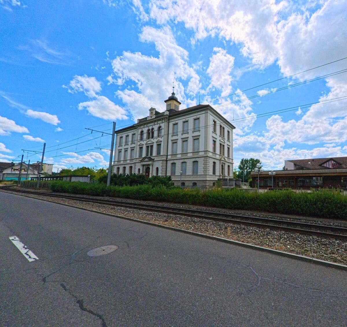 In Muhen (5037, Kanton Aargau) steht ein repraesentatives historisches Gebaeude mit Turmaufsatz direkt an der Bahnlinie, umgeben von Gruenflaechen und Gehwegen.