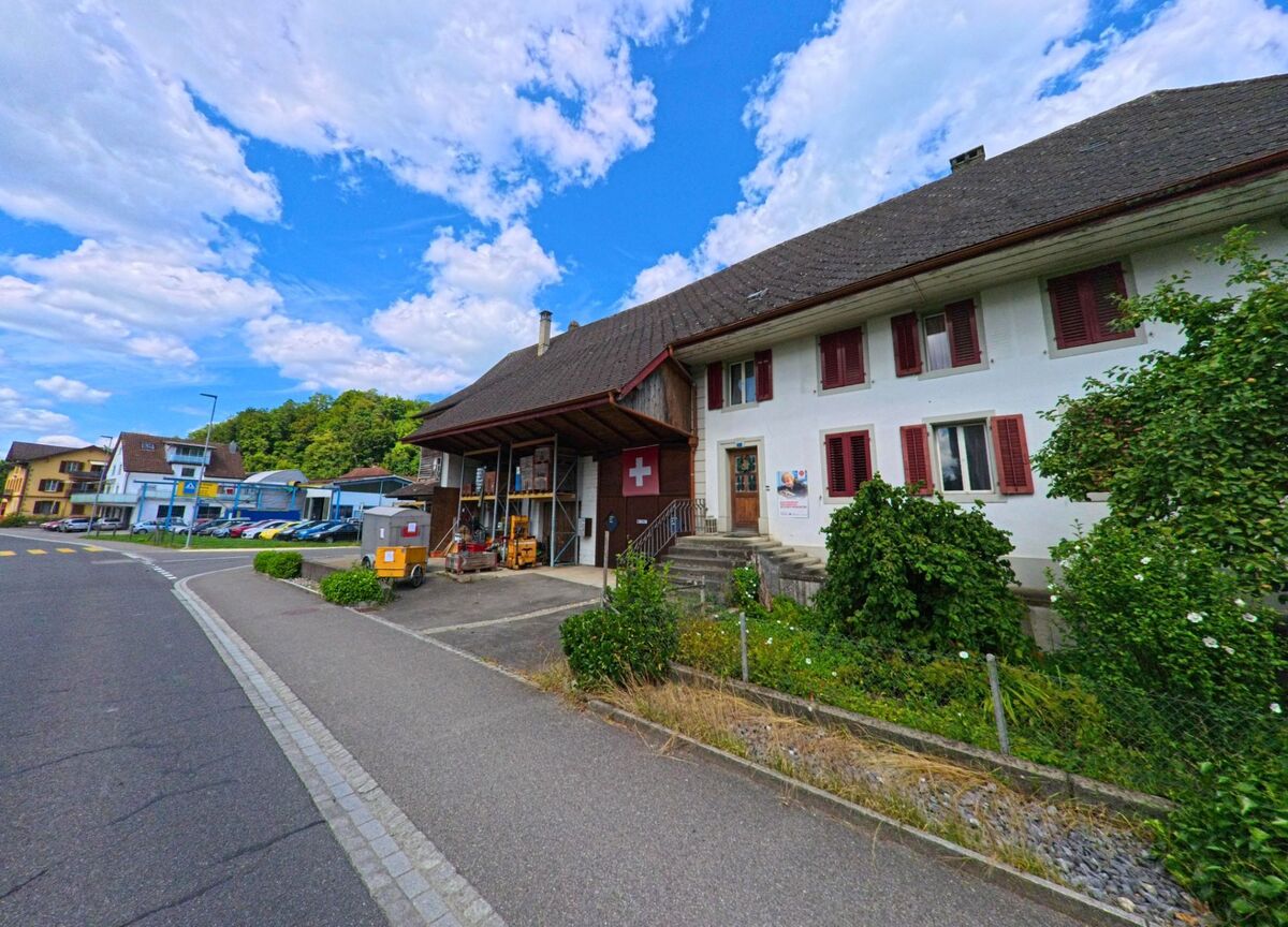 In Muhen (5037, Kanton Aargau) steht ein traditionelles Gebaeude mit rotem Fensterlaeden und angebautem Lagerbereich an einer leicht gekruemmten Strasse, im Hintergrund sind Geschaefte und Autogaragen sichtbar.