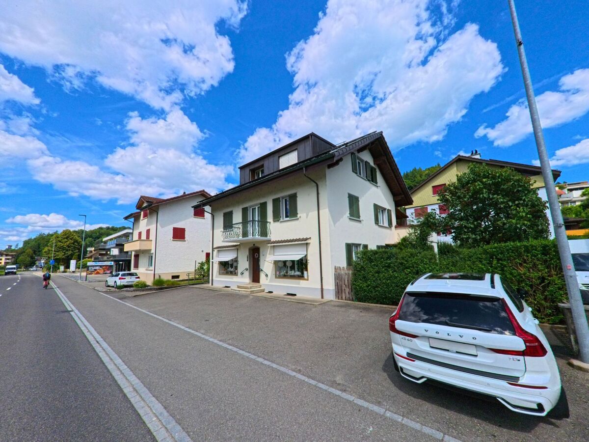 In Muhen (5037, Kanton Aargau) stehen entlang einer Hauptstrasse mehrere Ein- und Mehrfamilienhaeuser mit Vorgarten und Parkplaetzen, im Vordergrund ein weisses Fahrzeug.