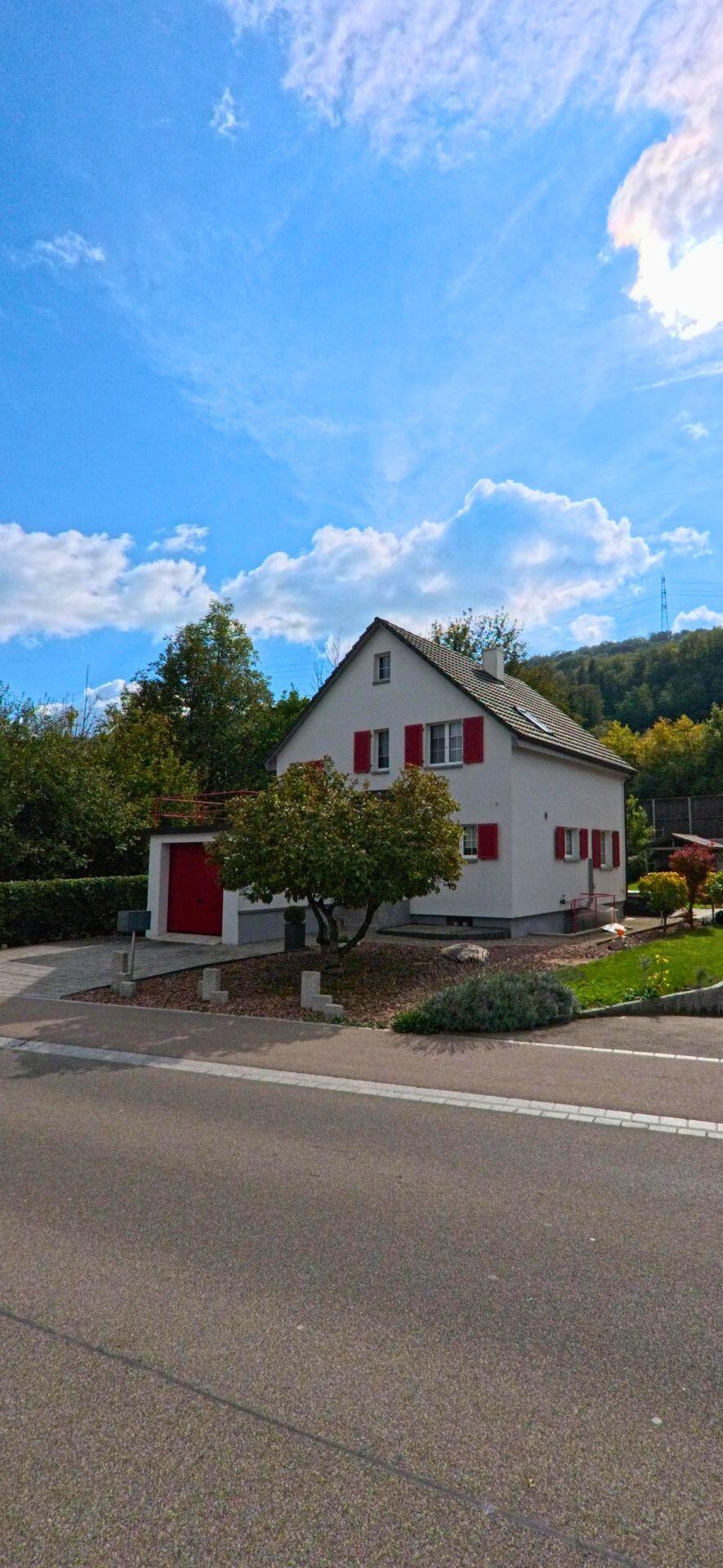 Ein Einfamilienhaus mit roten Fensterläden und gepflegtem Vorgarten steht an einer ruhigen Strasse in Mumpf (4322, Kanton Aargau) vor bewaldetem Hang.