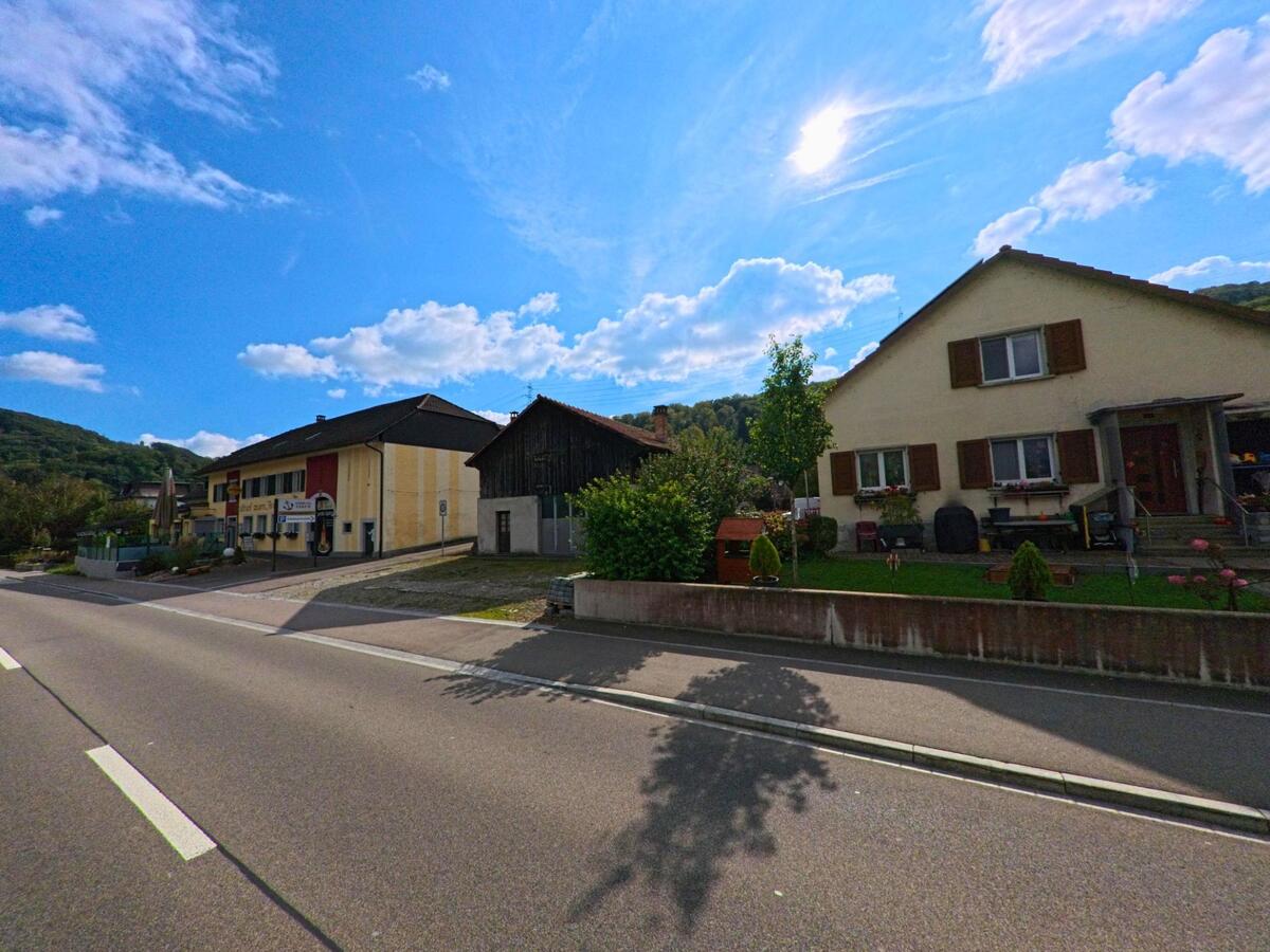 An der Hauptstrasse in Mumpf (4322, Kanton Aargau) stehen ein Wohnhaus mit Garten und ein gelbes Gasthaus mit Terrasse vor bewaldeten Hügeln.