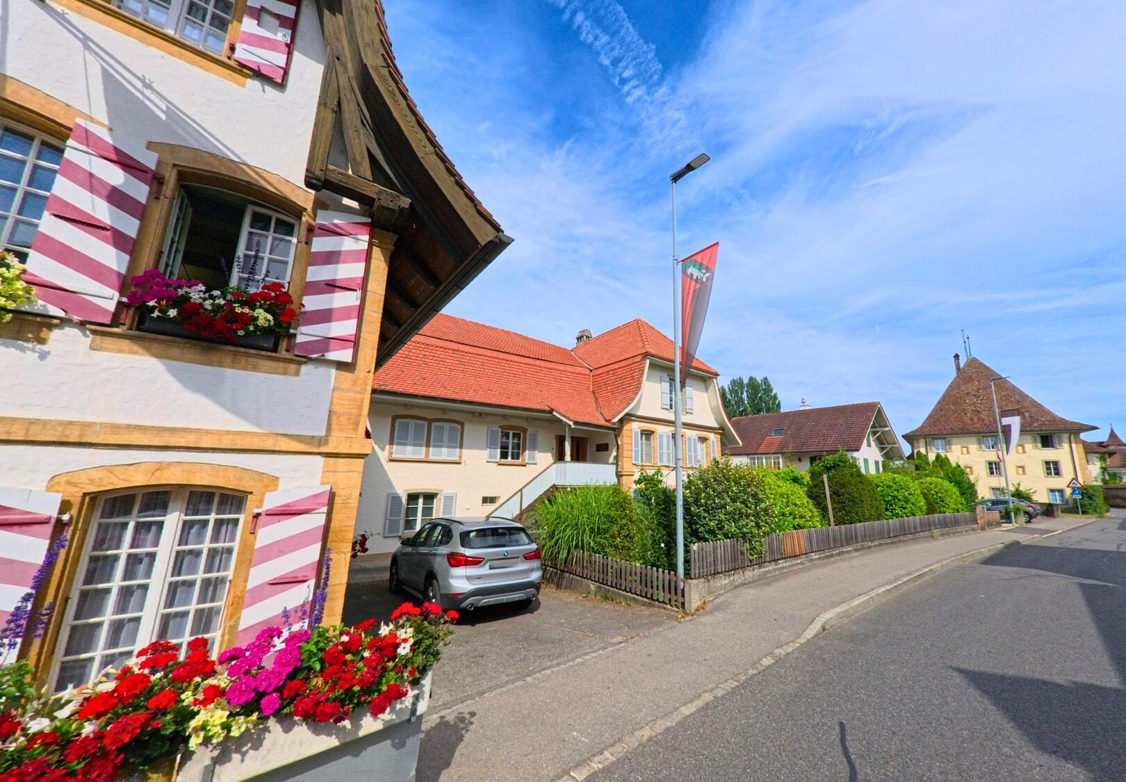 In Muntelier (3286, Kanton Freiburg) säumen blumengeschmückte Fenster und historische Gebäude mit roten Dächern eine ruhig verlaufende Dorfstrasse.