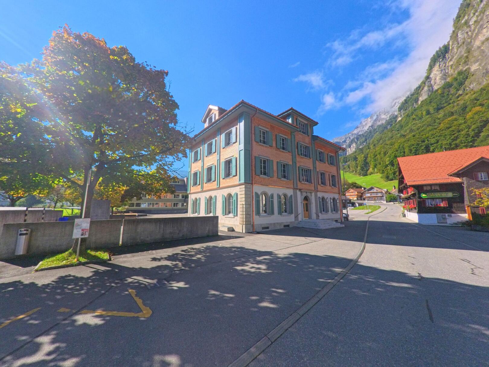 In Muotathal (6436, Kanton Schwyz) steht ein historisches Gebaeude mit gruenen Fenstern vor einer ansteigenden Strasse, die von alpinen Haengern und traditionellen Haeusern umgeben ist.