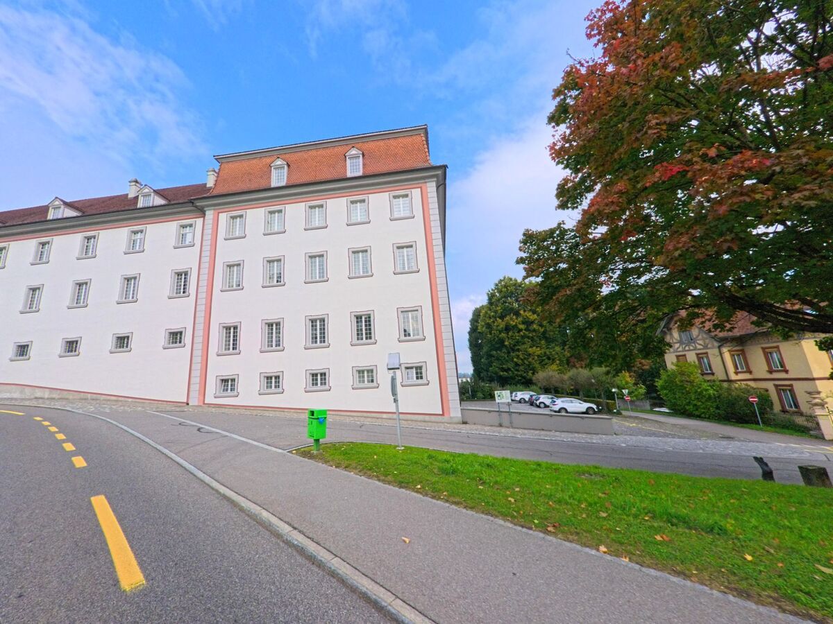 An einer leicht ansteigenden Strasse in Muri (5630, Kanton Aargau) steht ein grosses, weiss gestrichenes Gebaeude mit vielen Fenstern neben einem herbstlich belaubten Baum.