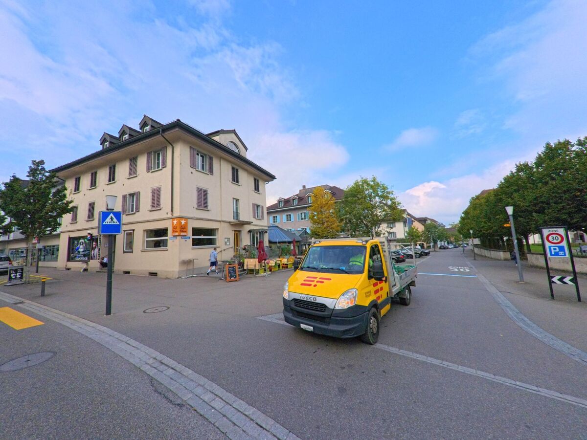 Im Zentrum von Muri (5630, Kanton Aargau) steht ein helles Wohn- und Geschaeftshaus an einer Kreuzung, wo ein gelbes Lieferfahrzeug vorbeifaehrt.