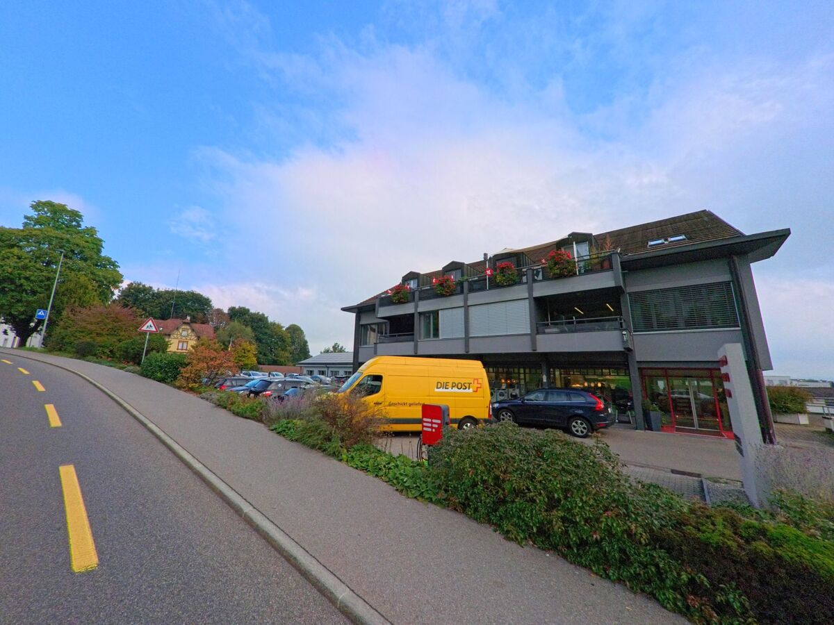 Ein modernes Gebaeude mit Balkonen und einem gelben Postauto steht an einer ansteigenden Strasse in Muri (5630, Kanton Aargau).