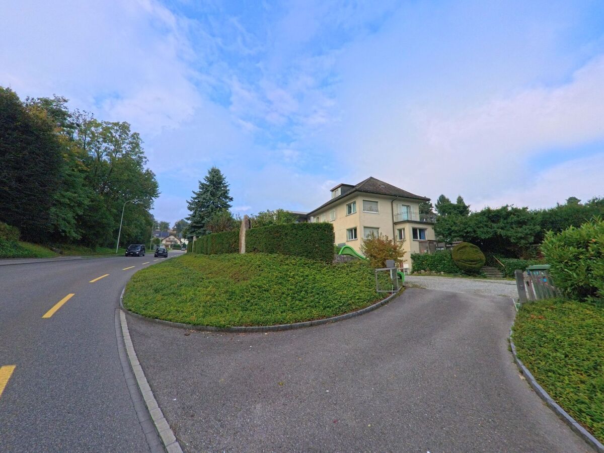 Ein helles Wohnhaus mit gepflegtem Garten liegt an einer leicht gekruemmten Strasse in Muri (5630, Kanton Aargau), umgeben von Baeumen und Gruenflaechen.