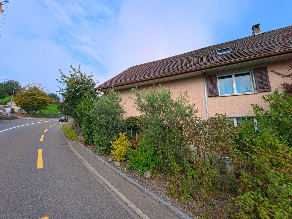 Ein Einfamilienhaus mit Garten und braunem Ziegeldach liegt an einer sanft gekruemmten Strasse in Muri (5630, Kanton Aargau).