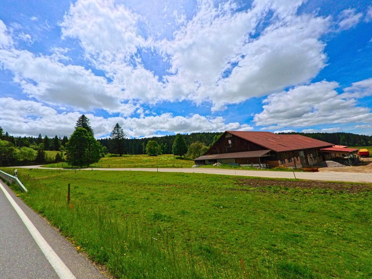 Eine ländliche Szene in Muriaux (2338, Kanton Jura) mit einer Straße, die an einem großen Bauernhof entlangführt, umgeben von Wiesen und Wäldern unter einem weiten Himmel.